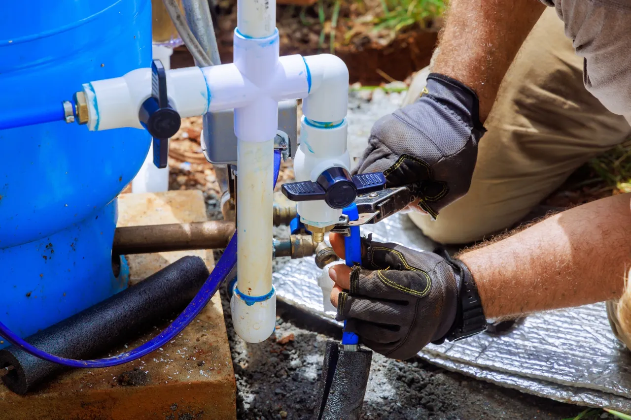 Técnico de luvas instalando registro e tubulação de PVC junto a tanque azul, etapa do custo de um poço